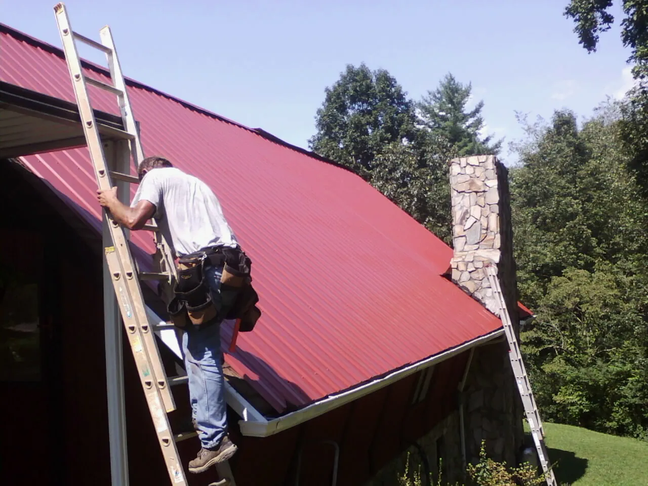 Roofer on a ladder beside a red metal roof and stone chimney — Soffit & Fascia Repair by Roofing Brothers in East Bethany